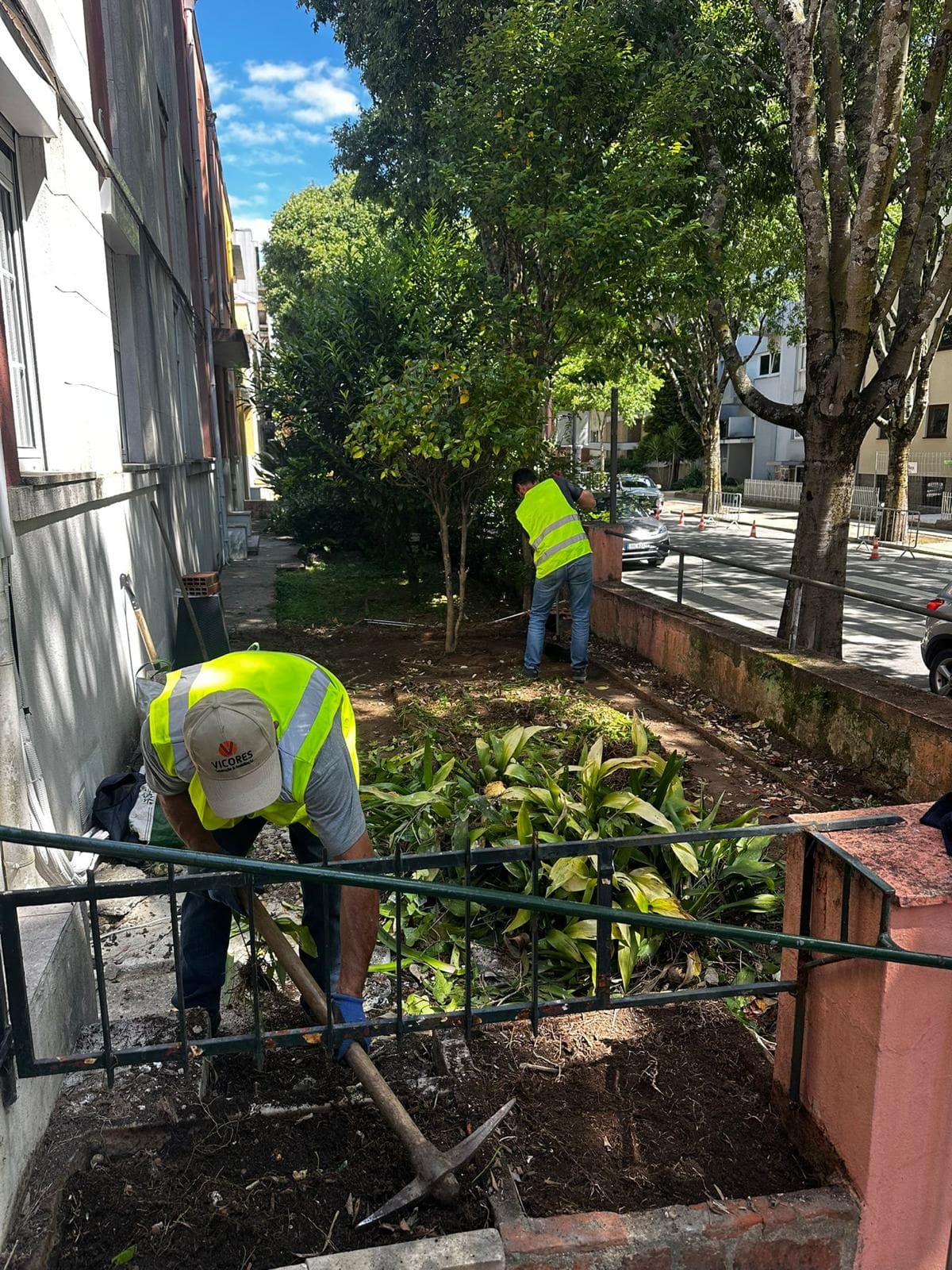 Trabalhador usa picareta enquanto outro limpa jardim urbano junto a um edifício e rua.