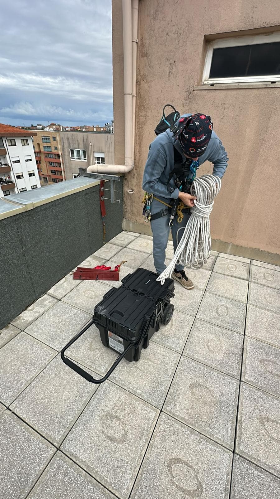 Trabalhador com arnês organiza cordas brancas e equipamentos de segurança no topo de um prédio.