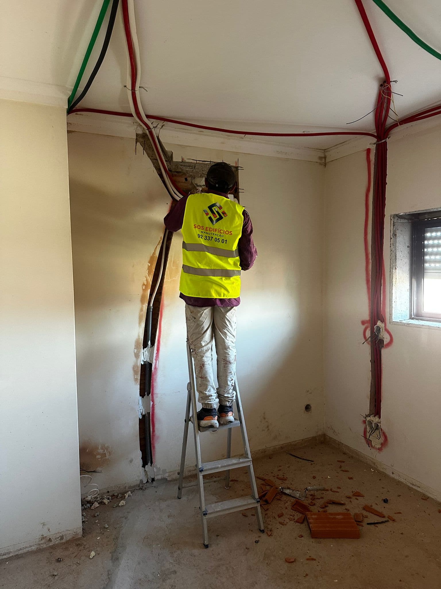 Trabalhador em escada instalando cabos elétricos em parede aberta durante reforma de ambiente interno.
